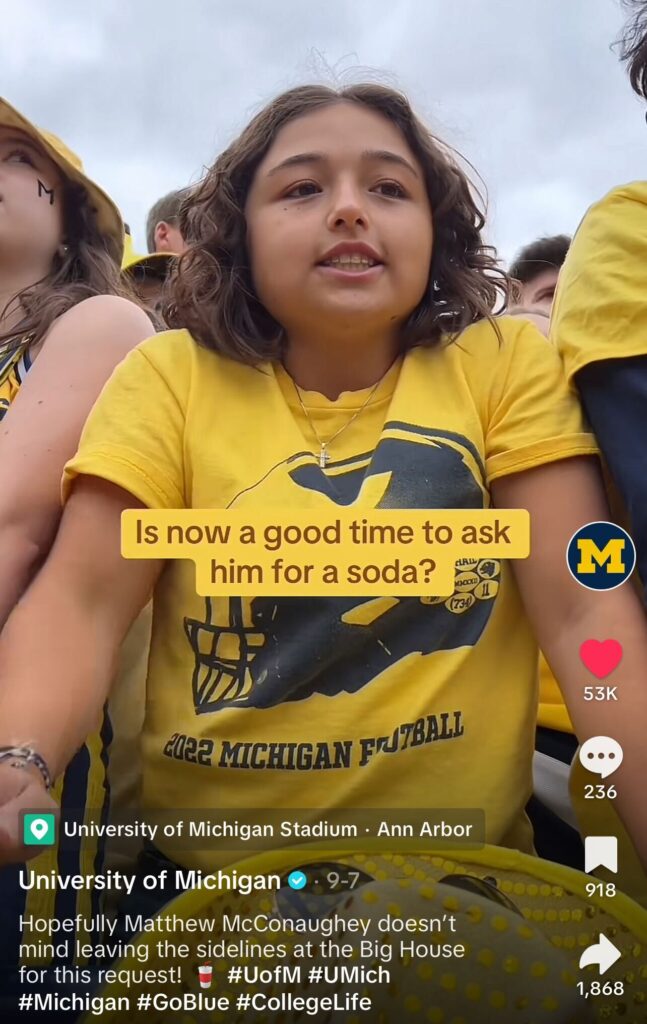 A screenshot of a TikTok showing a Michigan student at a Michigan football game with the text on screen reading "Is now a good time to ask him for a soda?" and the caption reading: Hopefully Matthew McConaughey doesn't mind leaving the sidelines at the Big House for this request!"